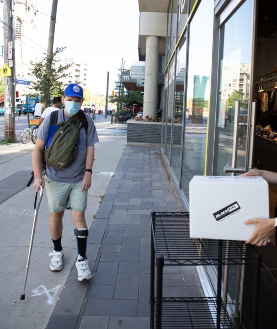A courier walking towards the store front where an individual is holding a package for him to collect.